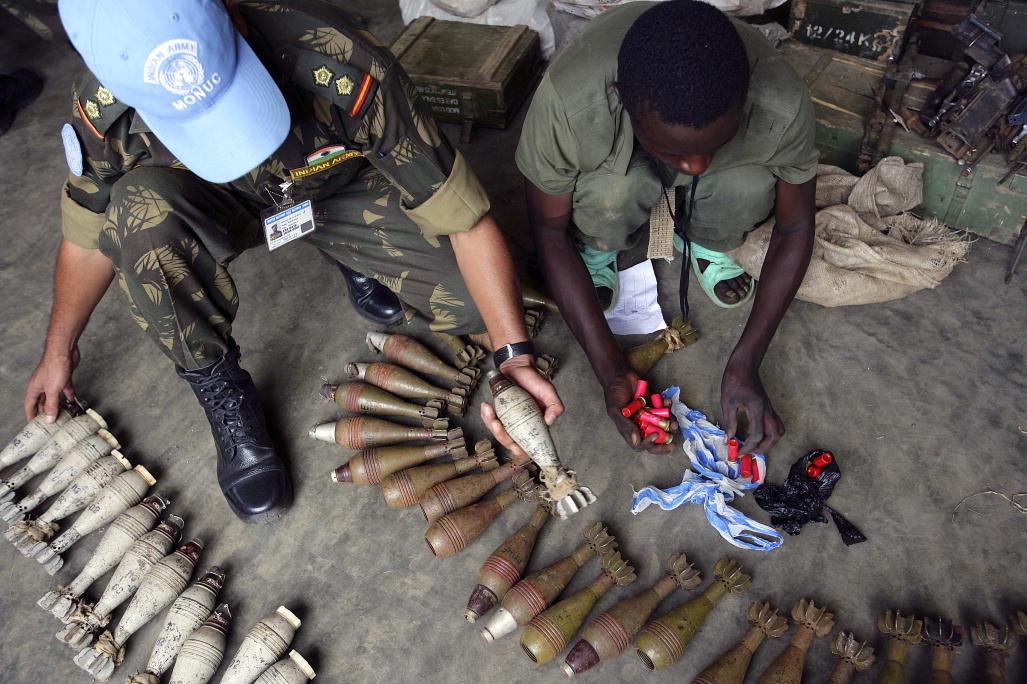 A man in fatigues and a blue UN ball cap kneels over and takes stock of arms while another man without a hat helps.