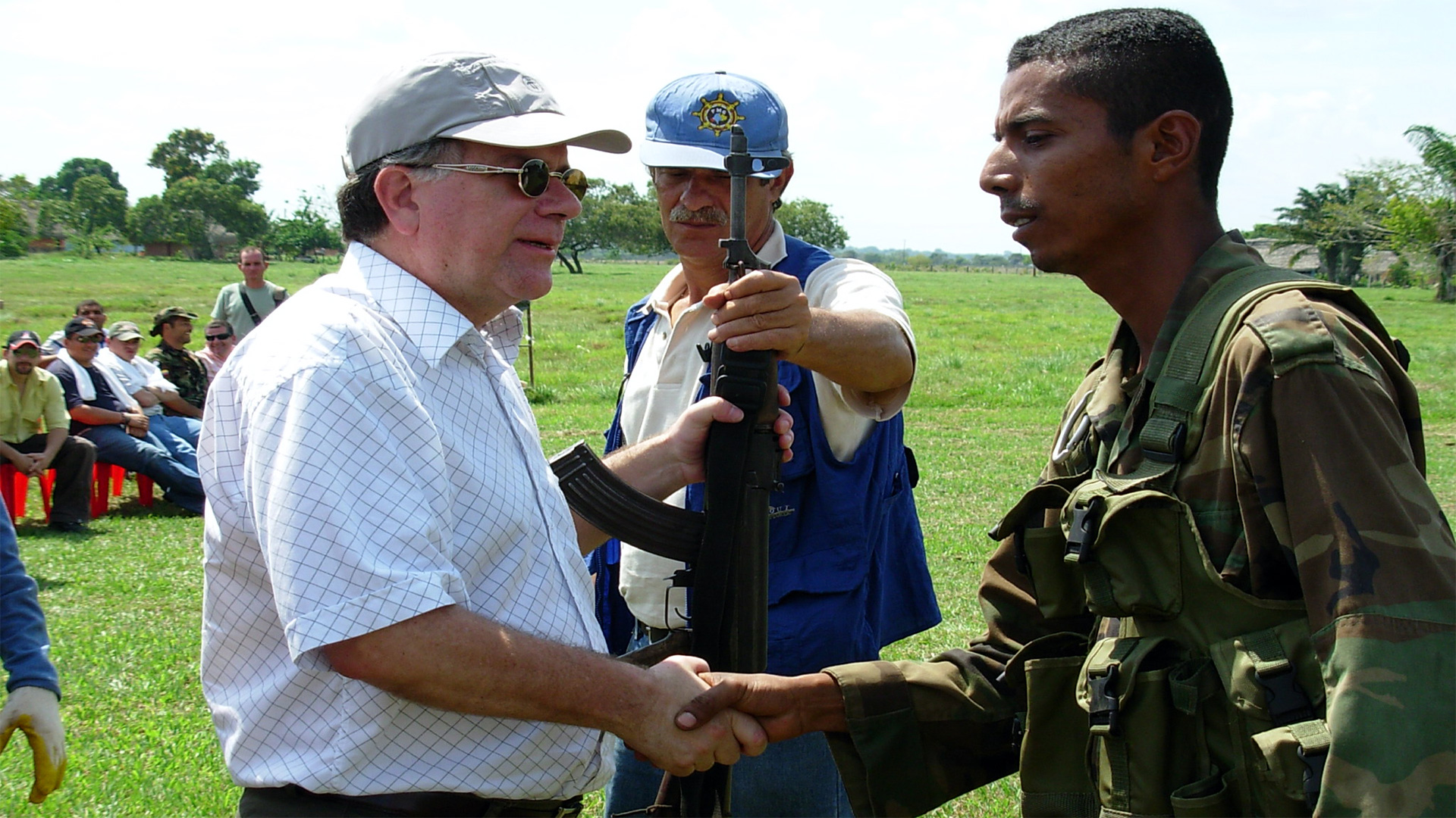 Two men shake hands in the field. One of them symbolically holds a rifle along with a third man