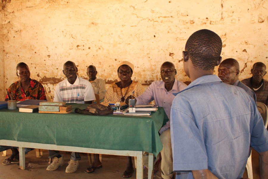 A man stands facing a table around which several men are seated.