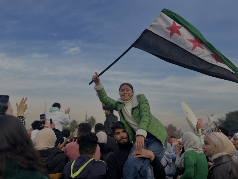 A large crowd of people in the street, many waving the Syrian flag