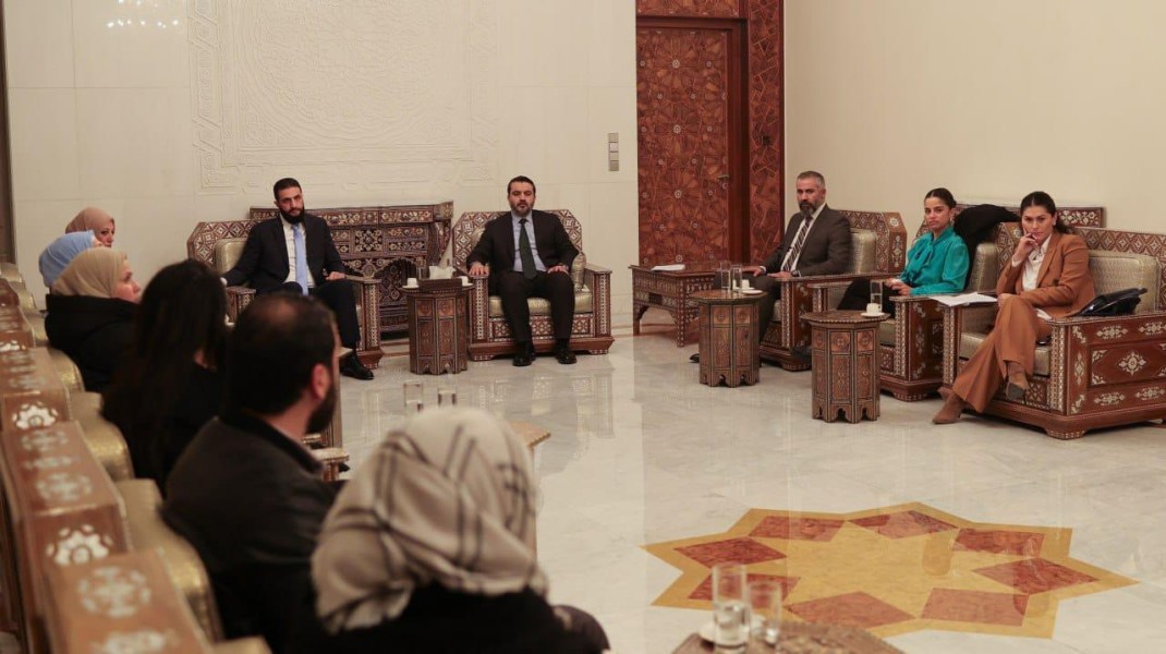Two men are seated at the front of a large elegant room. Another 10 or so people are seated in chairs along the side walls.