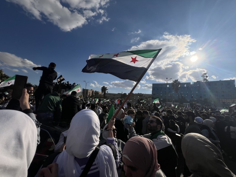 Large crowd of people in a city street some holding and waving the Syrian flags