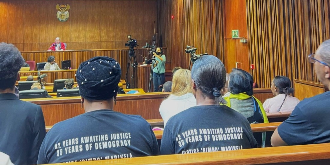Several people wearing T-shirts seated with backs camera in a courtroom and facing toward the judge who seated in an elevated stand and is facing the camera and audience