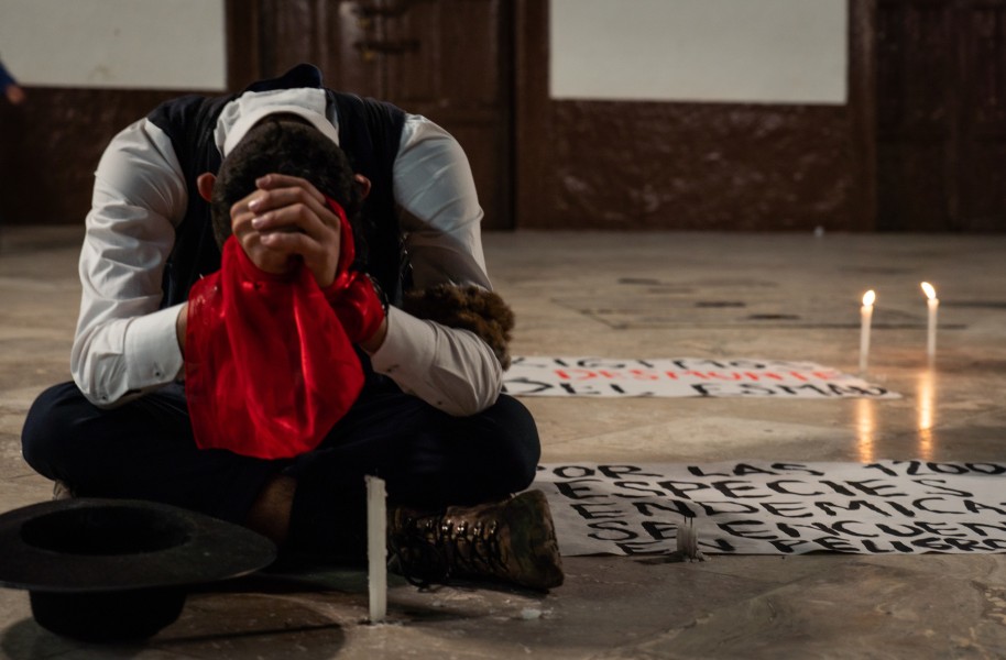 A man sits crossed legged with his head down. Next time to him are several lit candles.