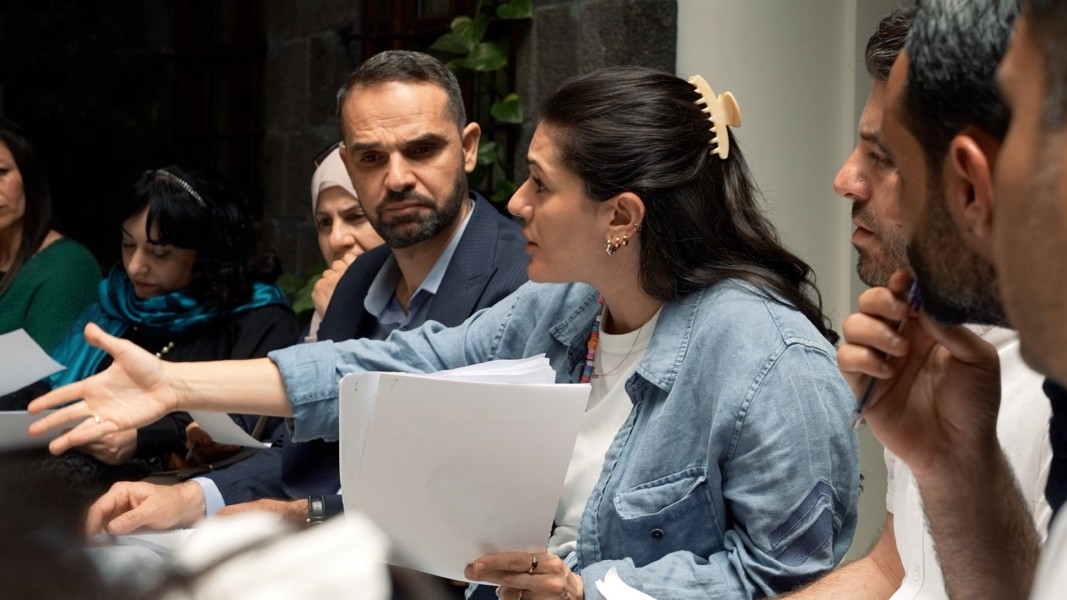 A women seated a table where other are also sitting is speaking; she is reaching out with one arm and holding papers in the other hand.