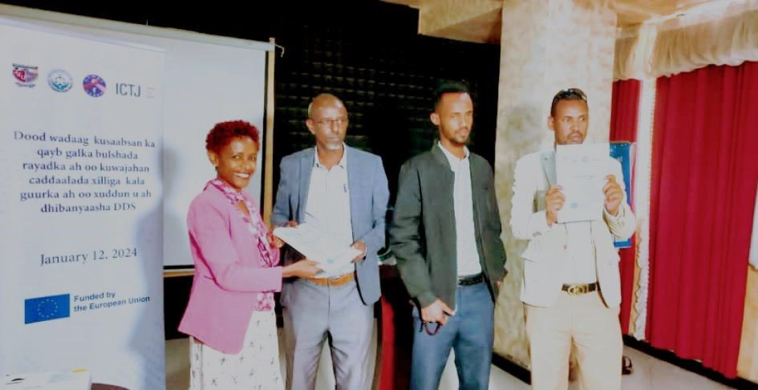 Four individuals stand in a room and next to a banner facing the camera and smiling; two of them hold a publication