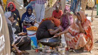 several women in colorful dresses crouch around an open fire pit, cooking food in a large pan.