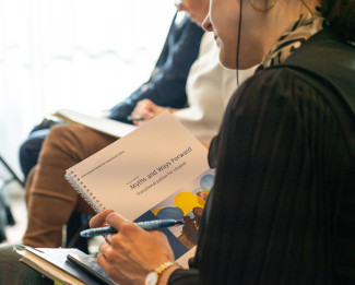 A seated person, back partially facing camera, holds a report.