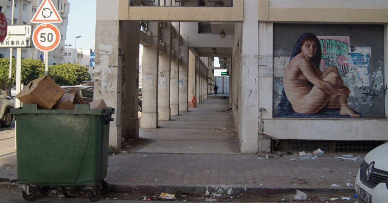 Color photograph of a city street and sidewalk, with a green dumpster on the left, and street art of a  woman in a blue robe on the right.