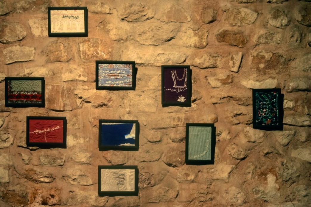 Nine black frames hang on a wall, each one framing a piece of fabric in a different color that has some embriodery on them.