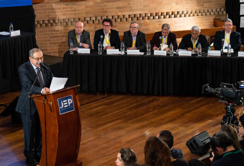 In a hearing chamber, a man addresses an audience at a podium, with six men seated behind him.