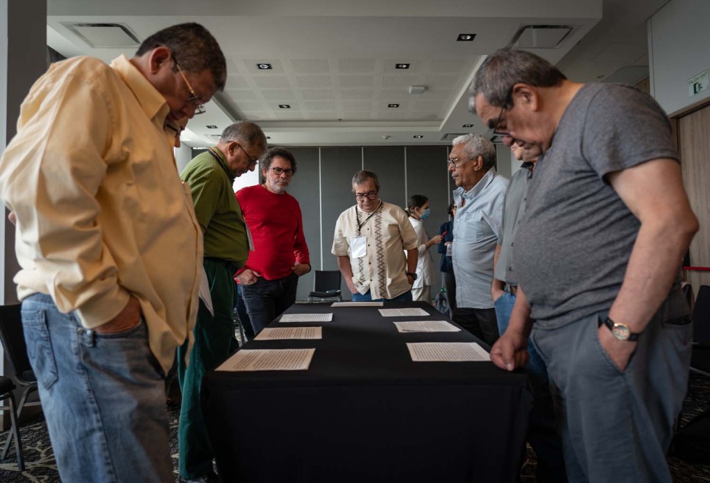 A group of seven man stand around a table, looking down at papers on the table top.