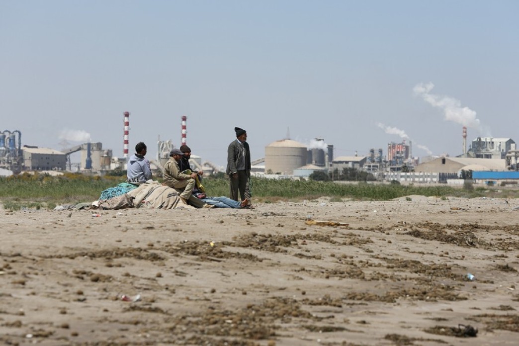 A group of fishermen stare towards the sea, the chemical plant looming behind them.